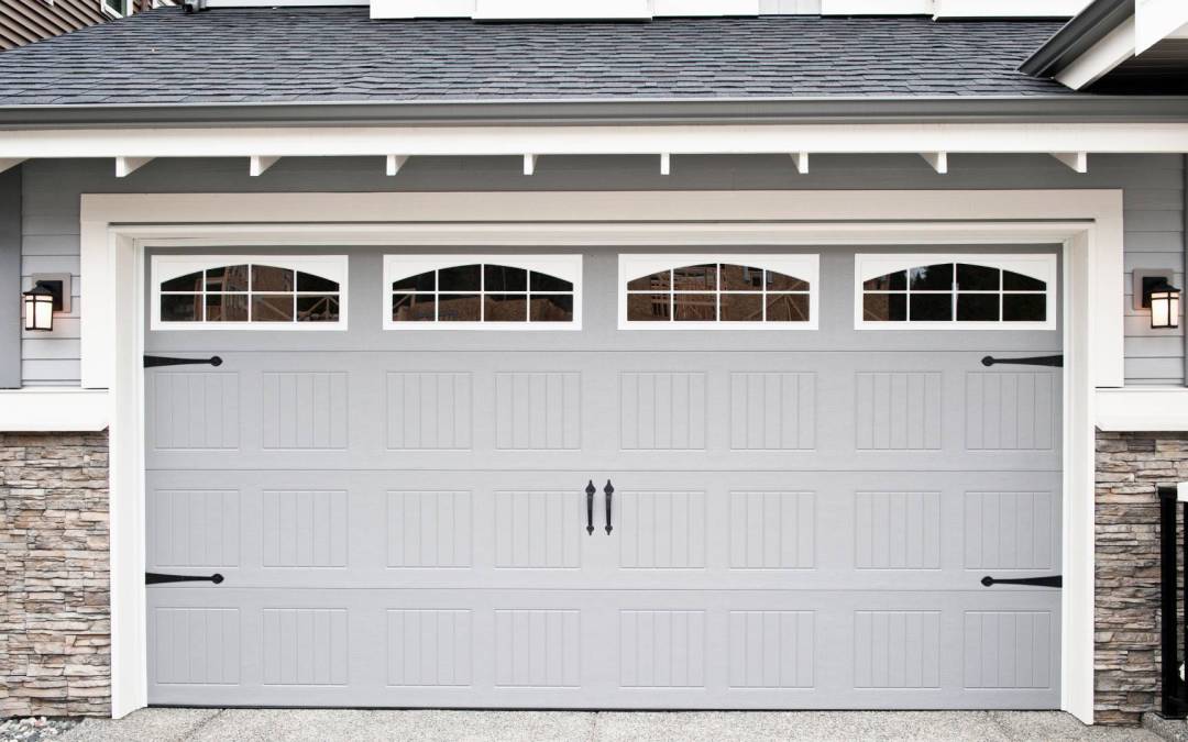 Front of a home’s garage with gray garage doors and black hardware, white wooden trim, black light fixtures, and a gray roof.