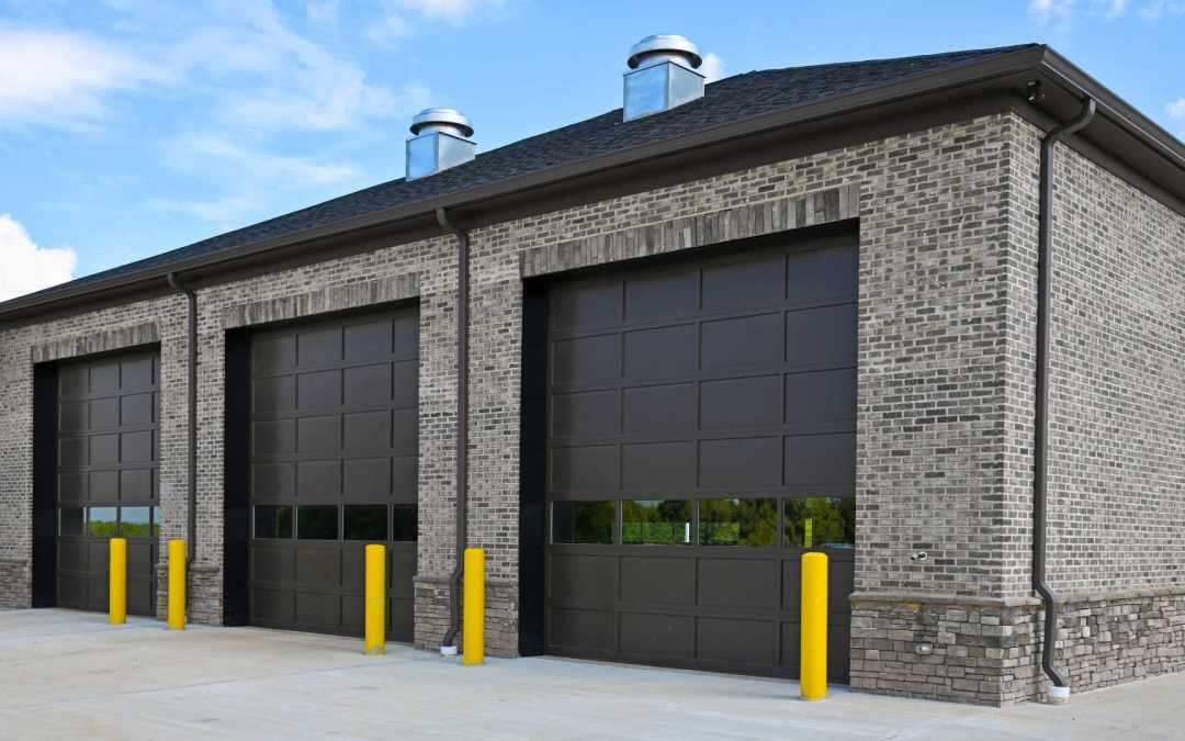 A large commercial garage made out of bricks has three large brown sectional garage doors and yellow safety poles.