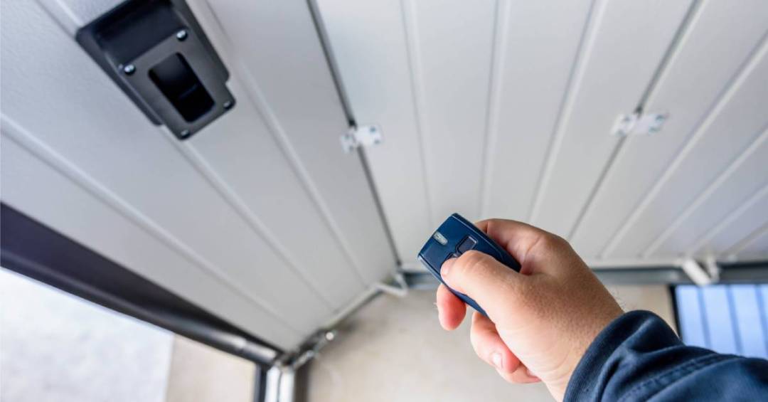 Close-up of a hand holding a small remote control to an opening white garage door with a sensor attached to the bottom.